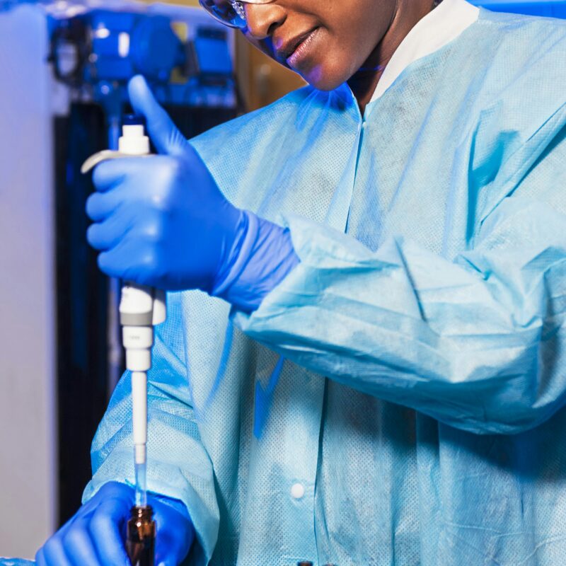 Une personne dans un laboratoire, utilisant une pipette pour ajouter des substances dans des tubes à essai, avec un équipement scientifique en arrière-plan.